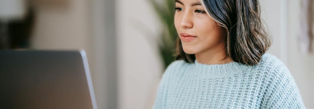 Confident young Latin American woman in casual clothes working remotely on netbook at table near mug with coffee in light apartment with plant with green leaves near wall